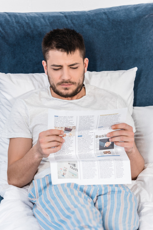 handsome man in pajamas reading newspaper in bed in morning at homeの写真素材