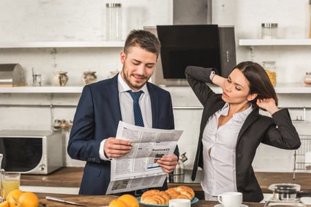 boyfriend reading newspaper and girlfriend touching hair in kitchen, gender equality conceptの写真素材