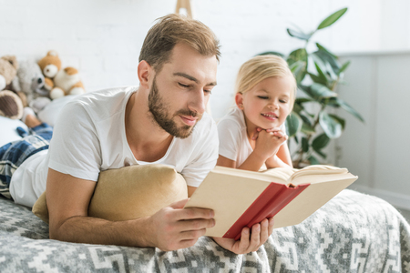 happy father and cute smiling daughter lying on bed and reading bookの写真素材