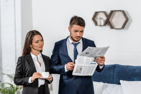 girlfriend and boyfriend in suits holding cups of coffee in morning, man reading newspaper at home, gender equality conceptの写真素材