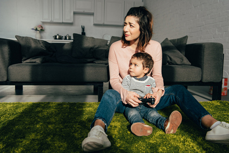 child and his mother sitting on floor and playing video game with joystick at homeの写真素材
