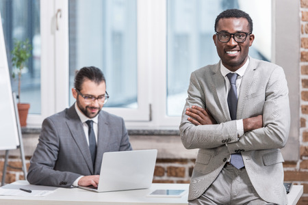 african american businessman with arms crossed looking at camera with partner working on laptop at backgroundの写真素材