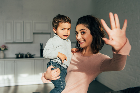 cheerful woman holding son and waving by hand at homeの写真素材