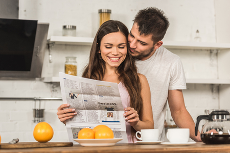 smiling girlfriend reading newspaper and boyfriend hugging her from back in morning at kitchenの写真素材
