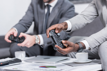 cropped view of multiethnic businessmen sitting at table with documents and holding gamepads