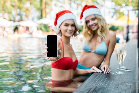 smiling women in swimming suits and santa claus hats showing smartphone with blank screen while relaxing in swimming poolの写真素材