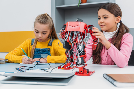 schoolgirls writing at notebook and touching red electric robot during stem lesson