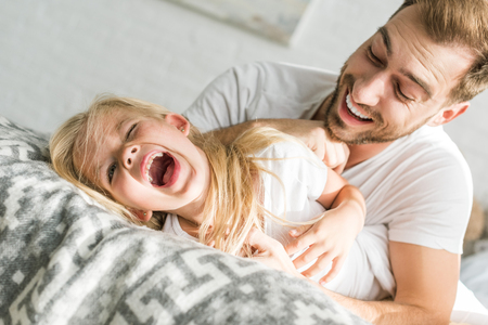 happy father tickling cute little daughter and having fun together in bedの写真素材