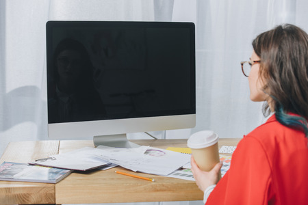 Young woman in glasses holding cup and working by computer