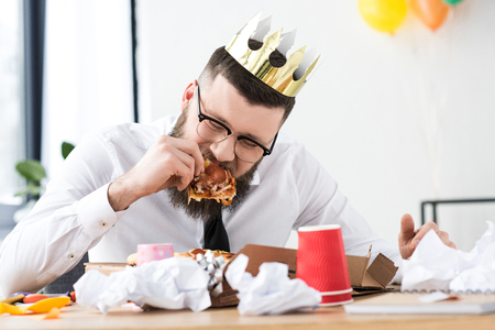 portrait of businessman with paper crown on head eating pizza at workplaceの写真素材
