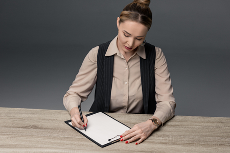 attractive kazakh businesswoman writing on clipboard at table isolated on greyの写真素材