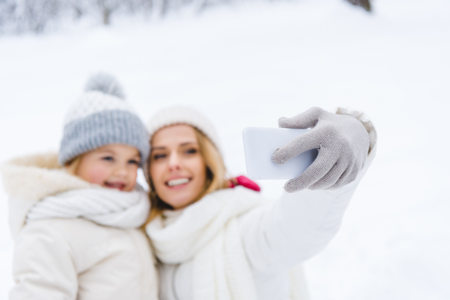 selective focus of happy mother and daughter taking selfie with smartphone in winter parkの写真素材