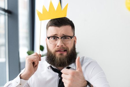 portrait of bearded businessman with paper crown showing thumb up in officeの写真素材