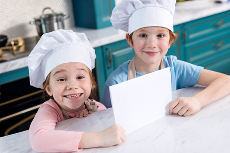 cute little children in chef hats holding blank card and smiling at cameraの写真素材