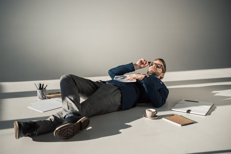 full length view of young businessman in eyeglasses holding newspaper and lying on floor