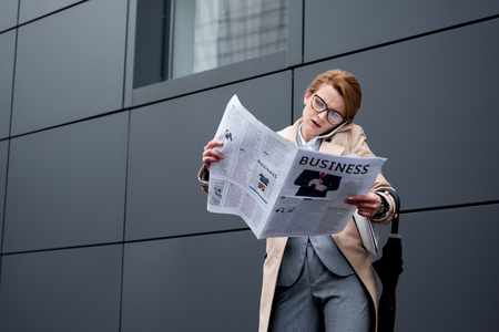 businesswoman talking on smartphone while reading newspaper on streetの写真素材