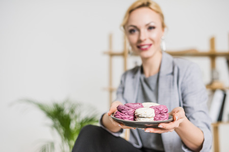 selective focus of smiling businesswoman holding plate with zephyr in hands in officeの写真素材