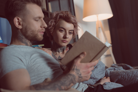 tattooed couple reading book while lying on sofa at homeの写真素材