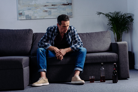 man looking at alcohol on floor while sitting on sofa at homeの写真素材