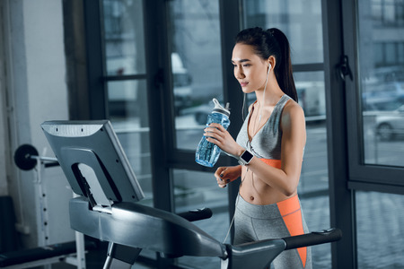 young athletic sportswoman drinking water while jogging on treadmill at gym