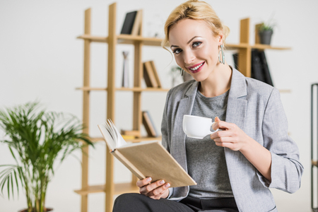 portrait of smiling businesswoman with book and cup of coffee in officeの写真素材