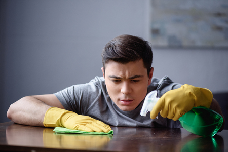 handsome man dusting table in living room with spray bottle and ragの写真素材