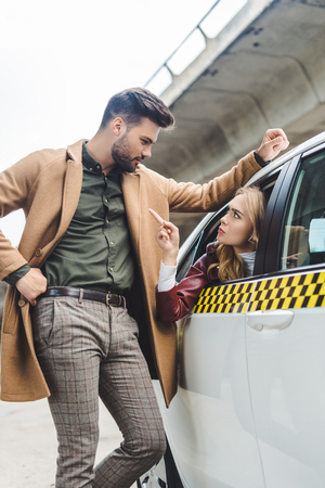 serious young woman sitting in taxi and pointing with finger at man leaning at carの写真素材