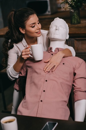 young woman with cup of coffee hugging mannequin at home, perfect relationship dream concept