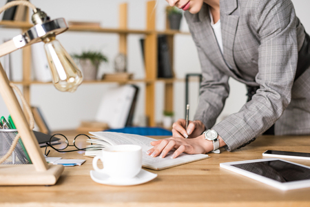 partial view of businesswoman making notes in notebook at workplace with cup of coffeeの写真素材