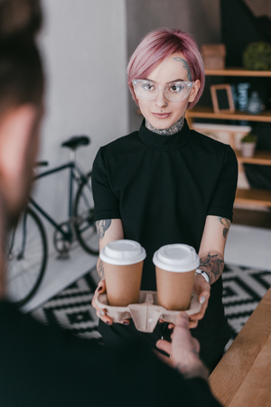 cropped shot of young tattooed woman giving paper cups to colleagueの写真素材