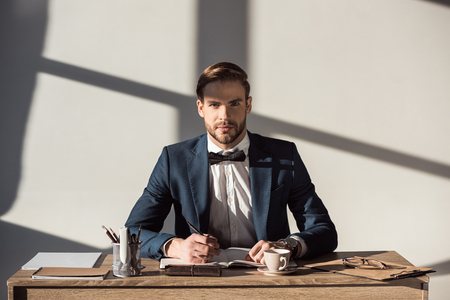 handsome stylish businessman looking at camera while writing in diary at workplaceの写真素材