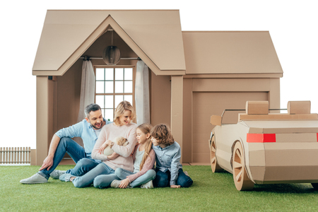 happy young family sitting on yard of cardboard house with their puppy isolated on whiteの写真素材