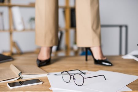 cropped shot of businesswoman standing near smartphone, papers and eyeglasses on floorの写真素材
