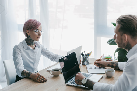 young businessman using laptop and looking at female colleague sitting oppositeの写真素材