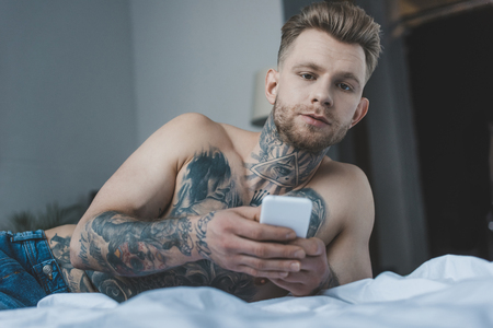 handsome shirtless tattooed man using smartphone on bedの写真素材