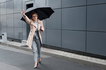 businesswoman in stylish coat with umbrella calling for taxi on streetの写真素材