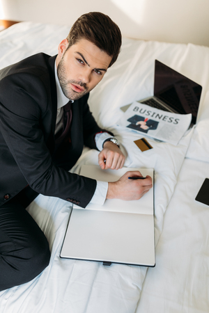 businessman lying on bed in hotel room with notebook and penの写真素材