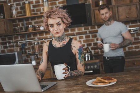 tattooed girl using laptop during breakfast, boyfriend behind in kitchenの写真素材