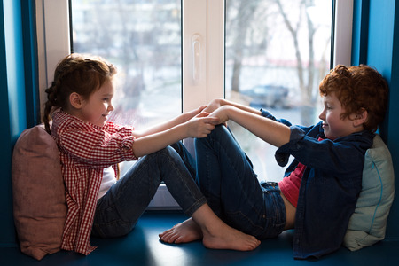 adorable little kids holding hands and smiling each other while sitting on windowsillの写真素材