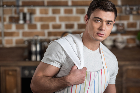 handsome man in apron with towel on shoulder looking at camera at kitchenの写真素材