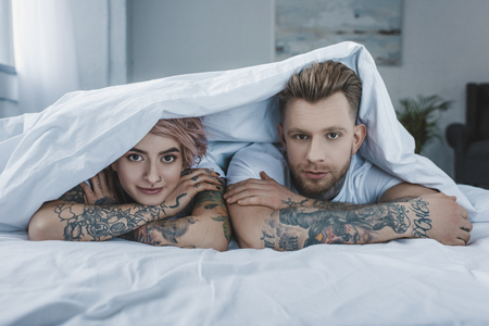 young tattooed couple lying in bed under white blanketの写真素材
