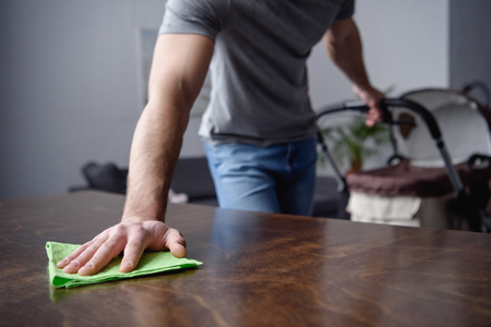 cropped image of man dusting table with rag and holding pram with other hand in living roomの写真素材