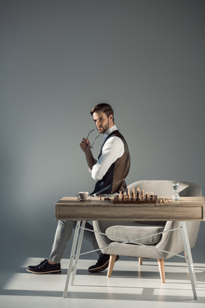 pensive stylish young businessman holding eyeglasses and looking away while playing chessの写真素材