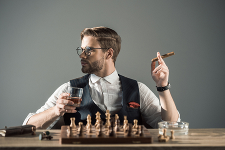 young man with glass of whisky smoking cigar and looking away while playing chessの写真素材