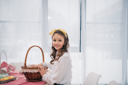 little child with basket looking at camera near table with easter eggs