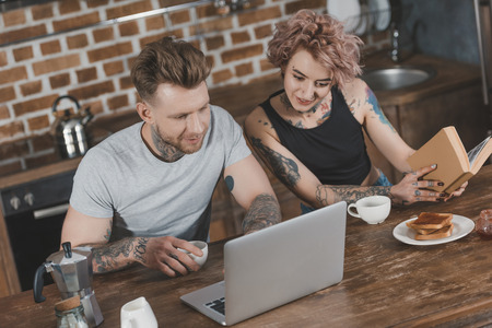 young couple using laptop and reading book during breakfast in kitchenの写真素材
