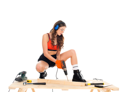 sexy girl in protective headphones working with electric drill at wooden table with tools, isolated on whiteの写真素材