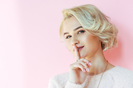 beautiful blonde girl gesturing for silence and looking at camera isolated on pinkの写真素材
