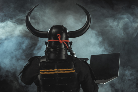 rear view of samurai in traditional armor with laptop on dark background with smokeの写真素材