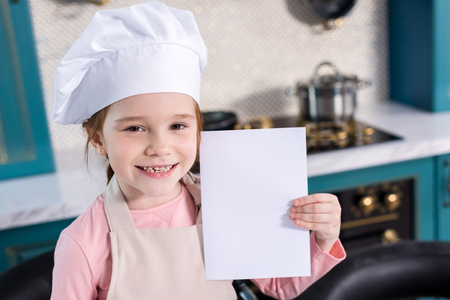 cute kid in chef hat and apron holding blank card and smiling at cameraの写真素材